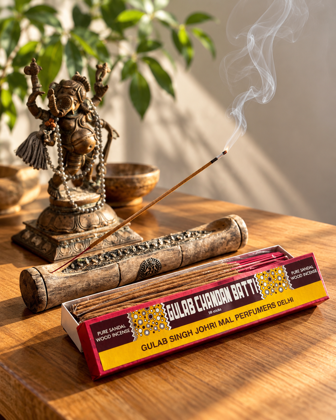 A box of Gulab Chandan Batti sandalwood incense open on a timber table, one stick burning in a carved holder beside a brass Ganesh statue, in warm afternoon light.